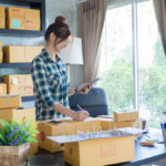 woman wearing plaid shirt with hair in bun and glasses packages items for delivery in her home office. packages are all around her on desk and shelving unit.