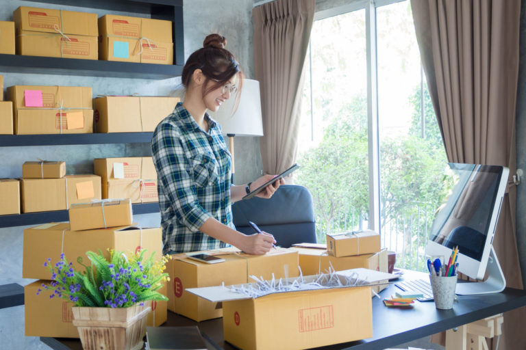 woman wearing plaid shirt with hair in bun and glasses packages items for delivery in her home office. packages are all around her on desk and shelving unit.