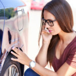 woman with long brown hair and glasses inspects the scratches on her car while talking on the phone