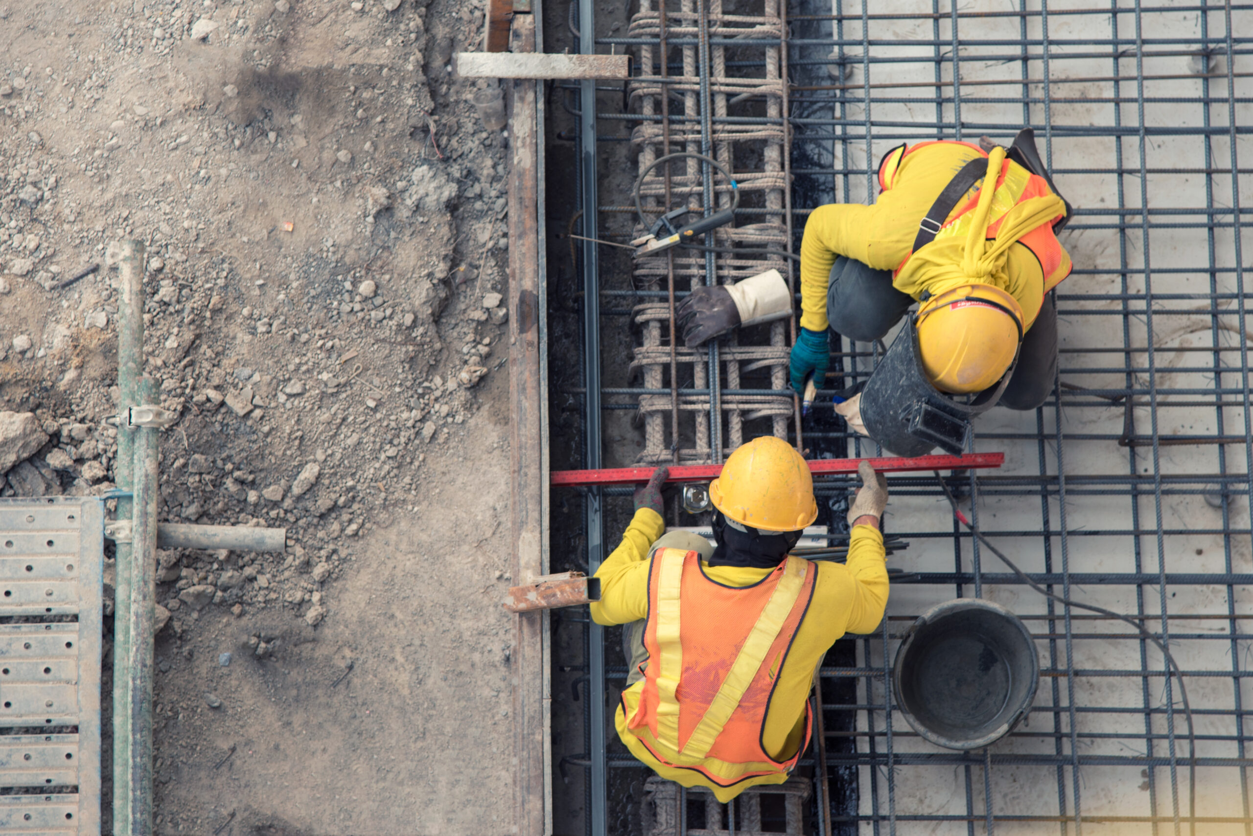 An overhead view of two construction workers installing steel rebar for a concrete foundation at an active job site. The image represents commercial construction, structural reinforcement, foundation work, and safe building practices.