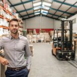 Man standing in a warehouse with inventory shelves and a forklift in the background